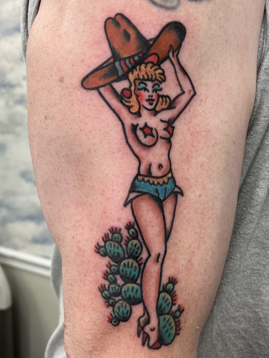 Cowgirl pin-up with arms raised under a big hat, standing among cactus on the upper arm, American Traditional tattoo by Chris at Diamond Mind Tattoo, Buzzards Bay
