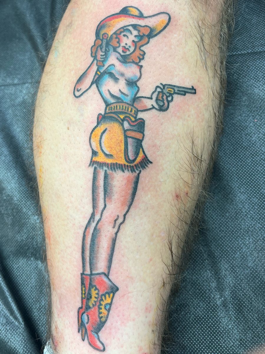 Cowgirl pin-up with a revolver, hat and red boots on the calf, American Traditional tattoo by Chris at Diamond Mind Tattoo, Buzzards Bay