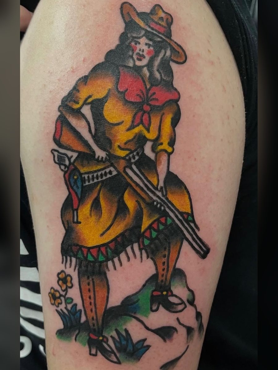 Cowgirl in a yellow fringed dress and hat with a rifle and holster, standing on a rock with flowers, on the upper arm, American Traditional tattoo by Chris at Diamond Mind Tattoo, Buzzards Bay