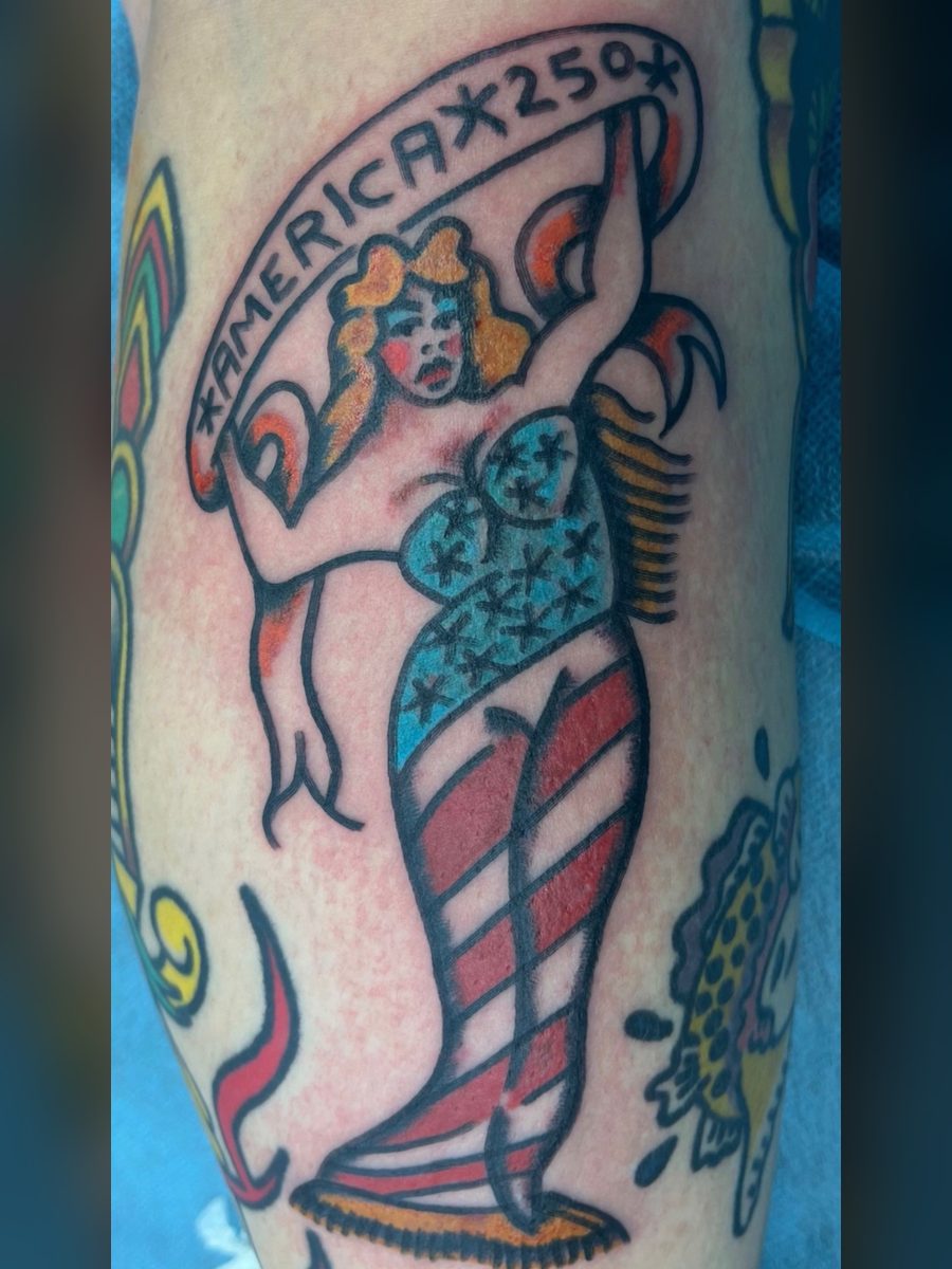 Pin-up woman in a stars-and-stripes dress holding an America 250 banner overhead on the leg, American Traditional tattoo by Chris at Diamond Mind Tattoo, Buzzards Bay