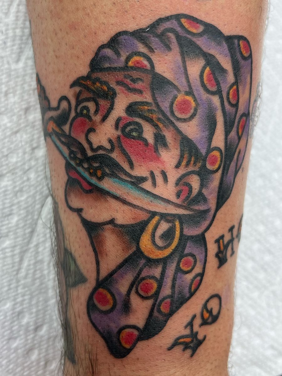 Pirate head in a purple polka-dot bandana with a knife in his teeth and a gold earring on the forearm, American Traditional tattoo by Chris at Diamond Mind Tattoo, Buzzards Bay